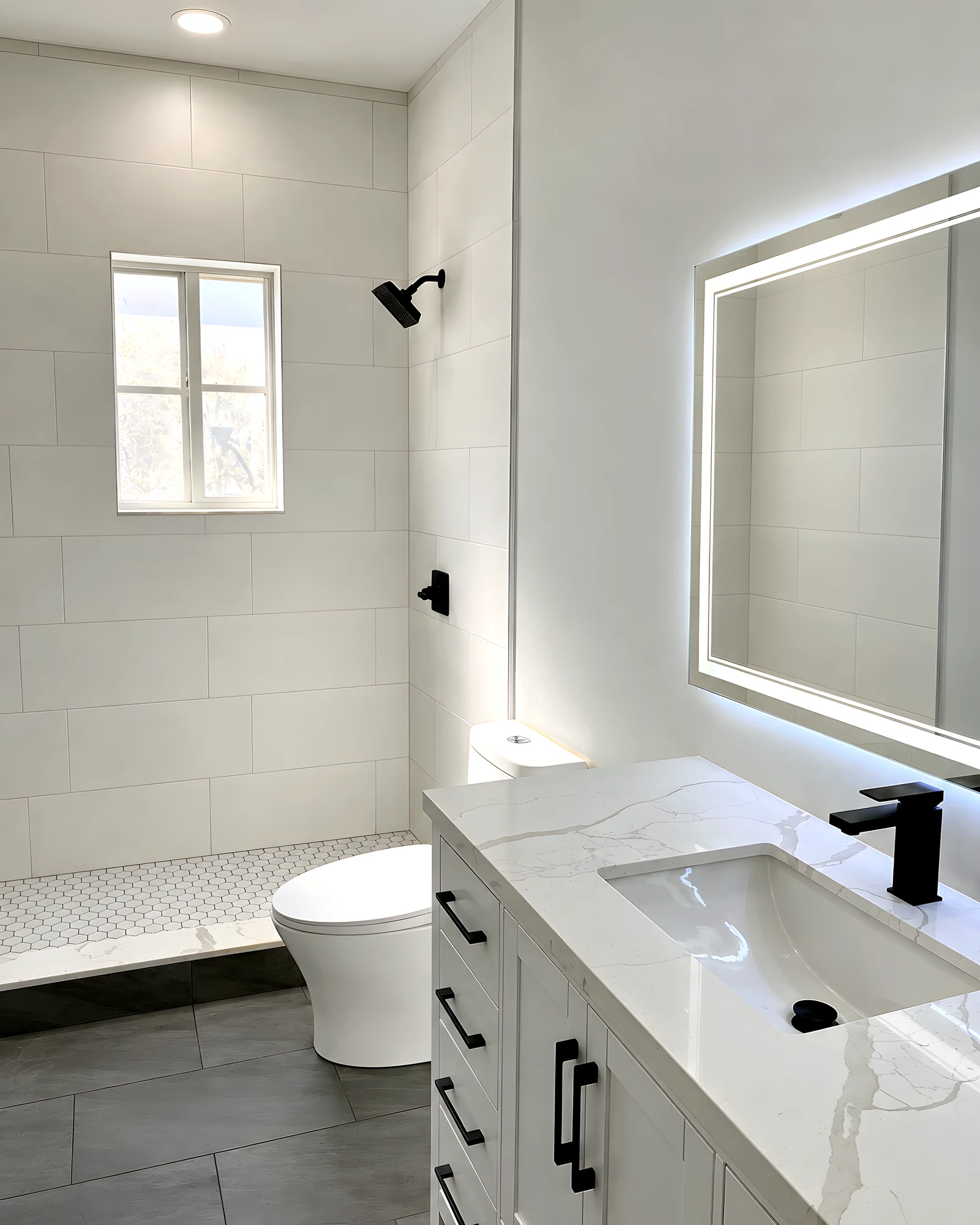 Baño con azulejo blanco de formato grande, ducha y grifería en negro mate, piso gris, mueble blanco con cuarzo y espejo LED